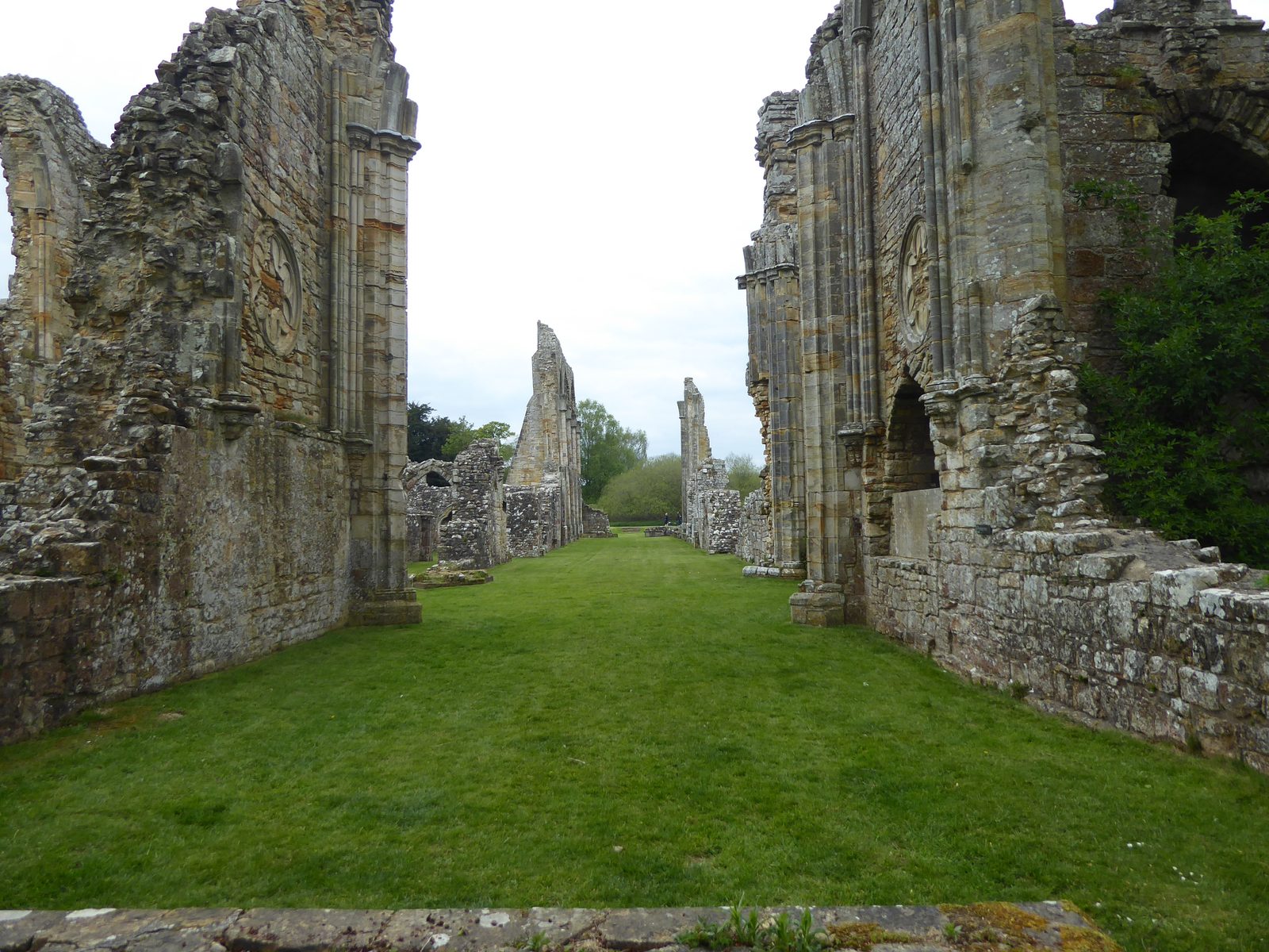 Looking down the roofless nave of the abbey church at Bayham Old Abbey, tall ruined arches either side and mown grass underfoot