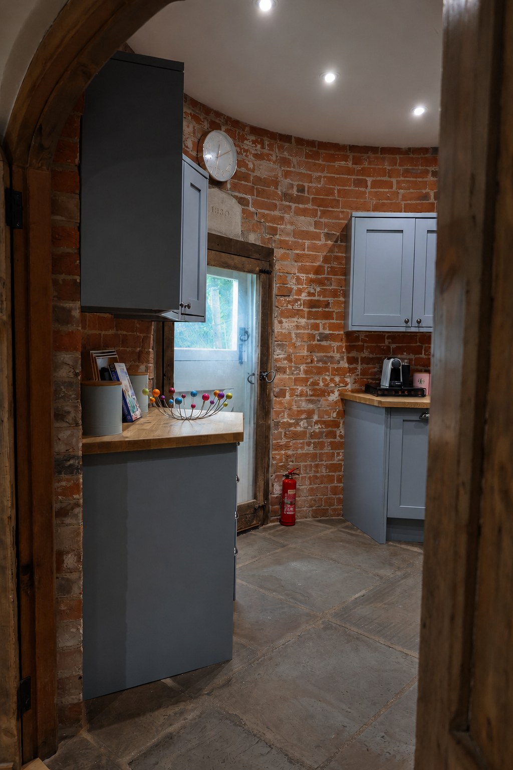 Looking through the timber arch towards the kitchen door at The Oast, with exposed brick, blue-grey cupboards and wooden worktops