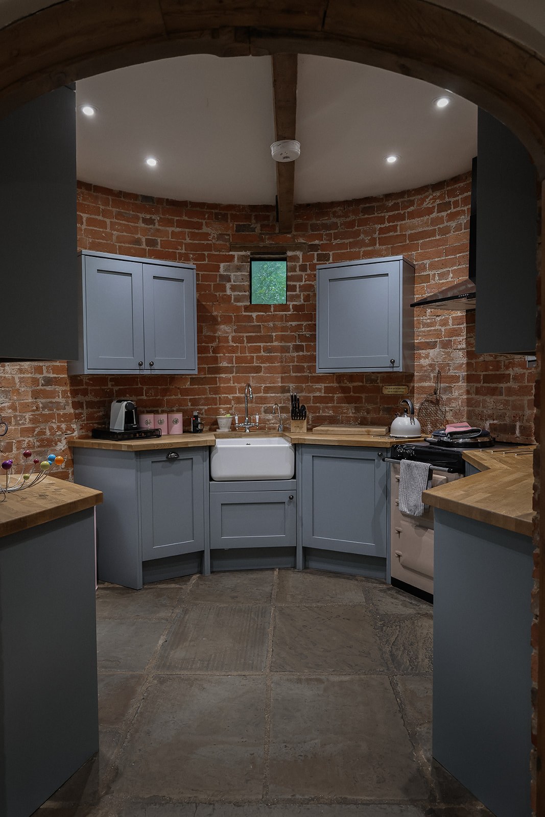 The round brick kitchen at The Oast, with blue-grey cupboards, a white sink, timber worktops and the pink AGA