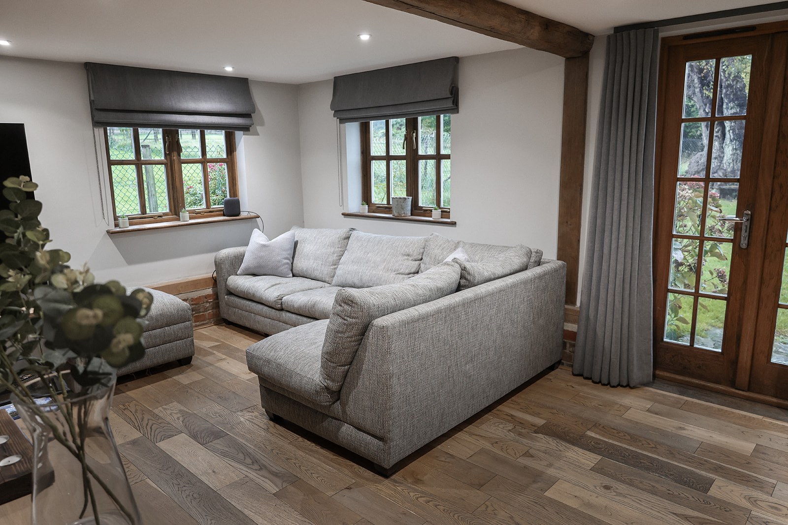 The sitting room at The Oast, with a grey corner sofa, timber beams, two windows and doors to the garden