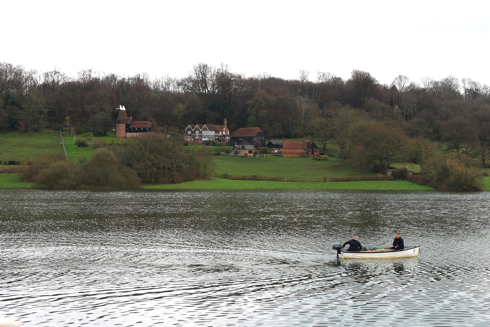 A small boat crossing the open water of Bewl reservoir, with wooded banks and a tile-hung farmhouse on the far shore