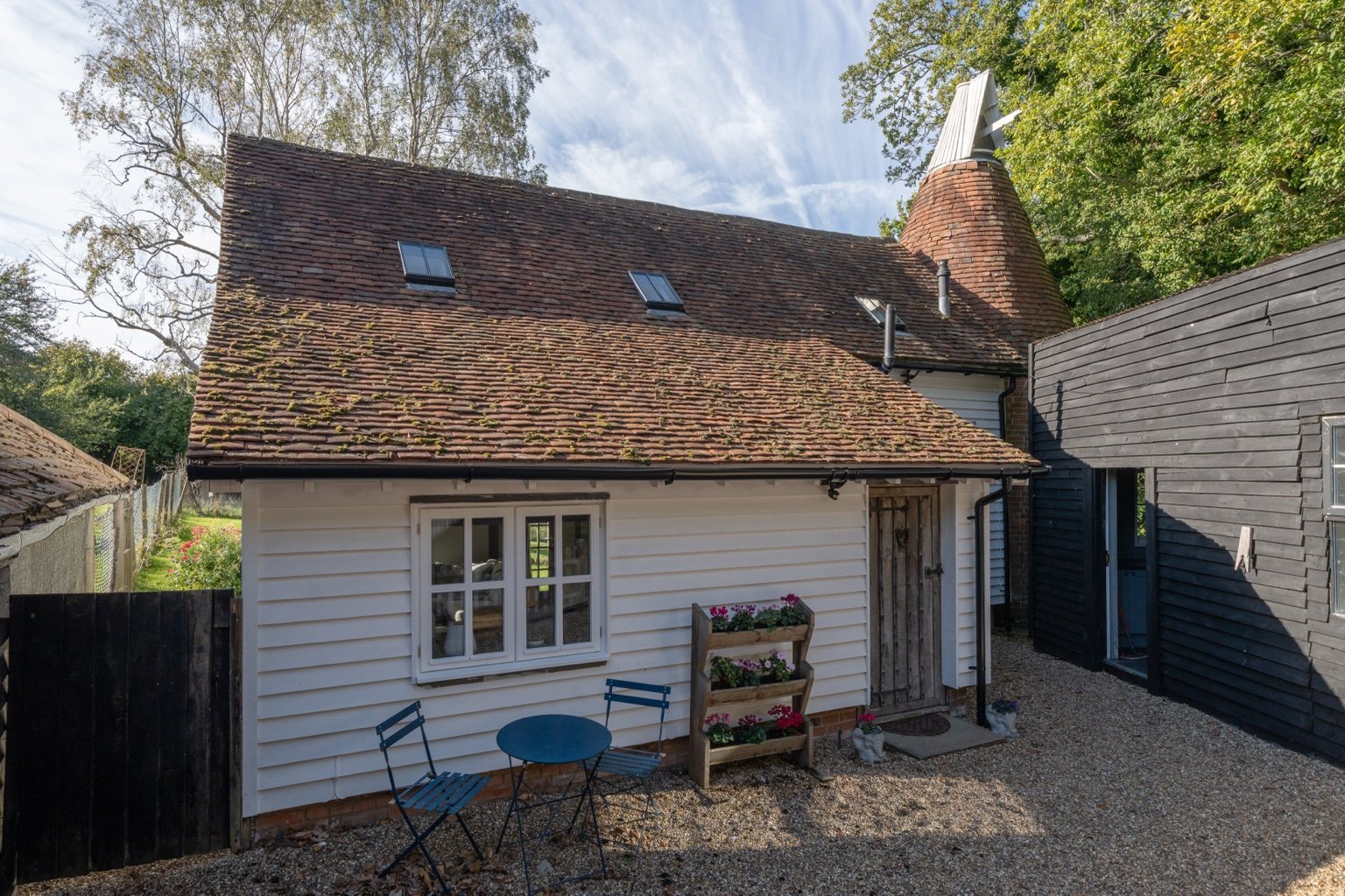 The courtyard entrance to The Oast, with white weatherboarding, tiled kiln roof and cowl, flowers and a blue table and chairs