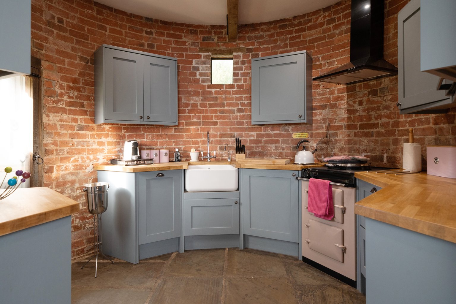 The round brick kitchen at The Oast, with grey cabinets, a white farmhouse sink, wooden worktops and the black AGA