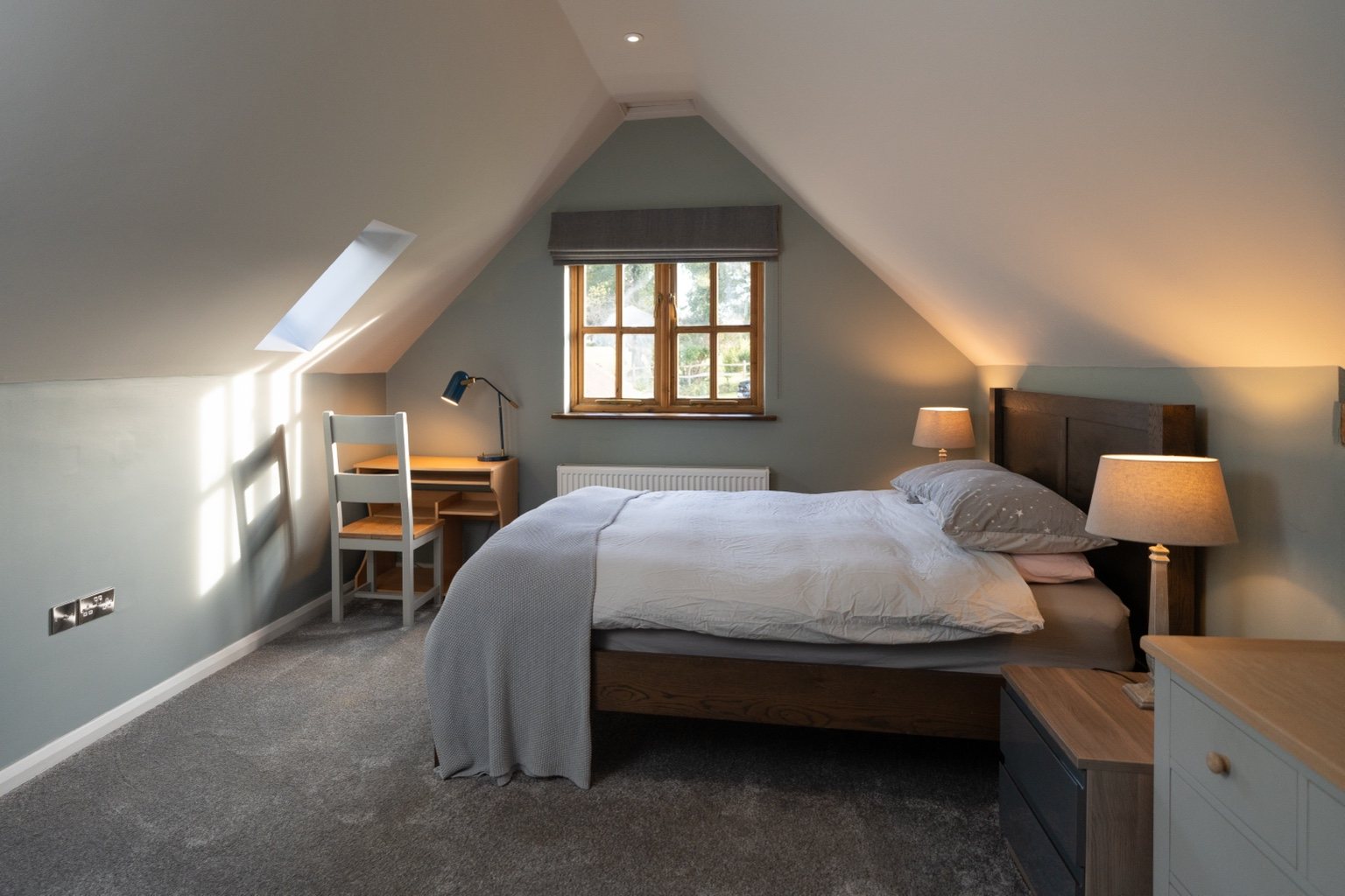The main bedroom at The Oast, with a wooden double bed, grey bedding, bedside lamps and a desk beneath the sloping ceiling