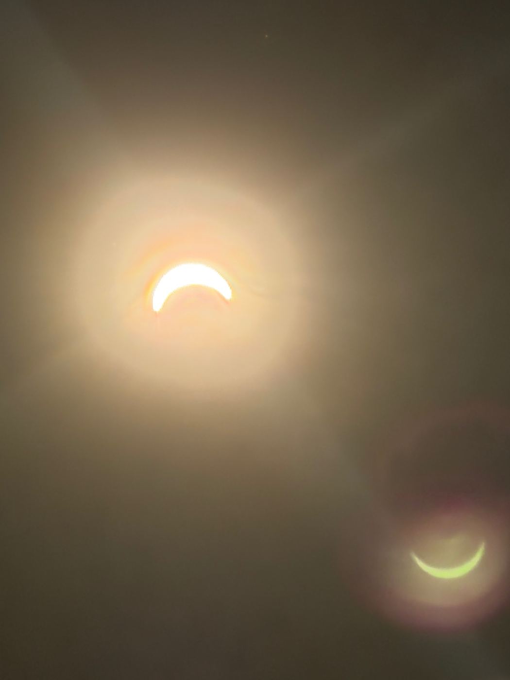 A close view of the eclipse at its deepest from Frant, the sun a thin white crescent in a soft amber glow, with a green crescent lens reflection in the lower right corner