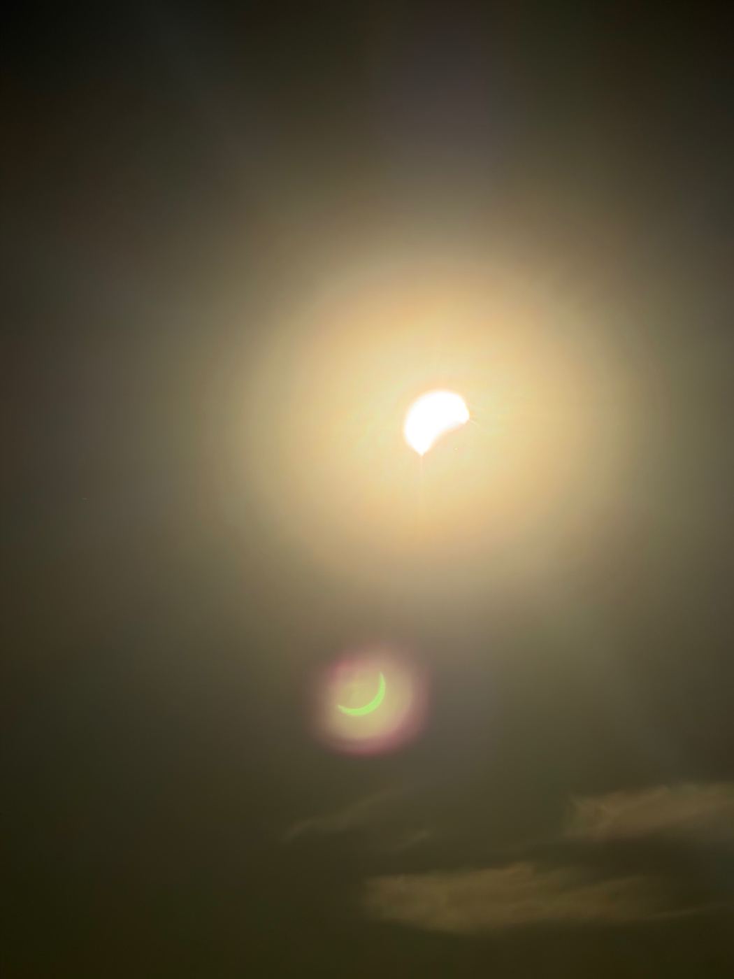 The eclipsed sun reduced to a bright crescent within a wide golden halo, with a small green crescent of lens flare mirroring it lower in the frame