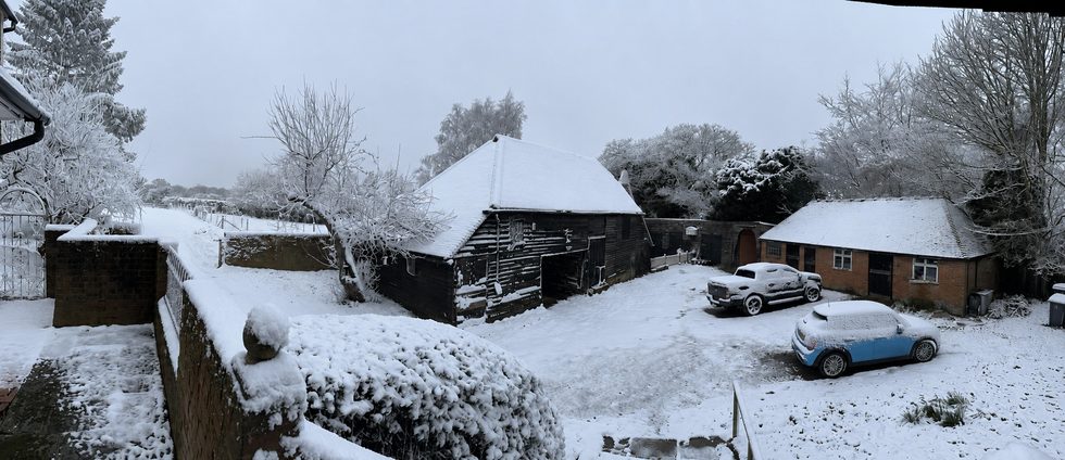 A panoramic view of Martins Farm under snow, with the black weatherboarded barn, brick walls, snow-covered vehicles in the yard and bare trees beyond