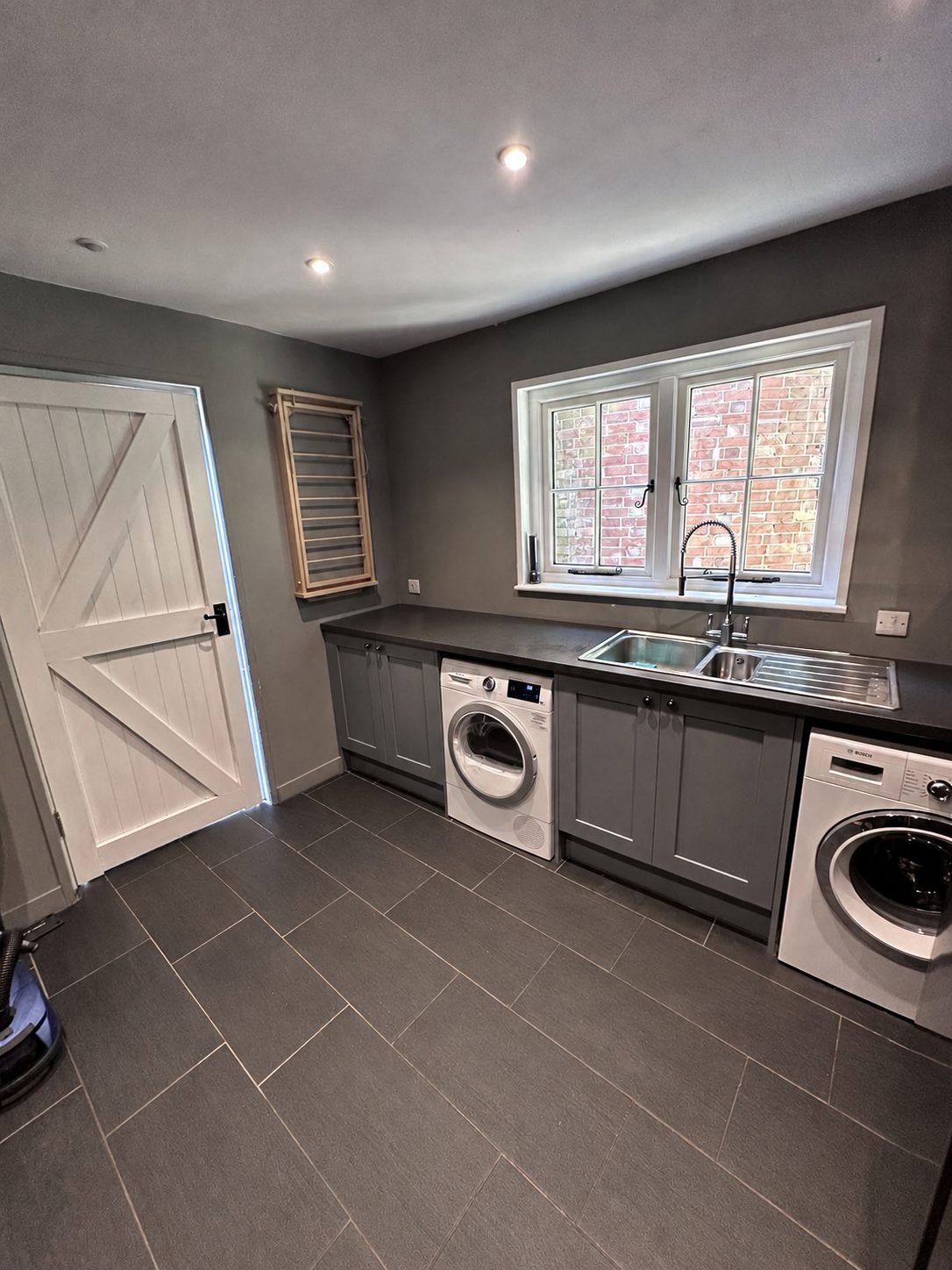 A boot room with grey cabinets, dark worktops, a double sink, a washing machine, a tumble dryer and a wooden drying rack on the wall