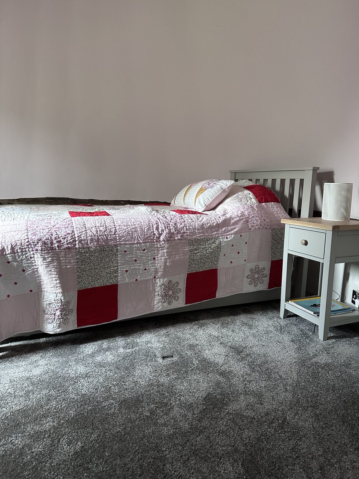 A single bed in a round bedroom at The Oast, made up with a pink and red patchwork quilt against a grey upholstered headboard, with a pale nightstand and white lamp beside it
