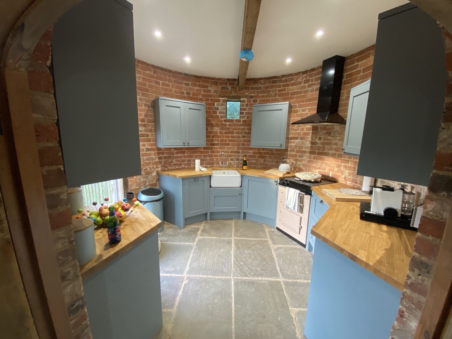 The kitchen in the round kiln at The Oast, with red brick walls, blue-grey cabinets, wooden worktops, a white farmhouse sink and a black AGA range