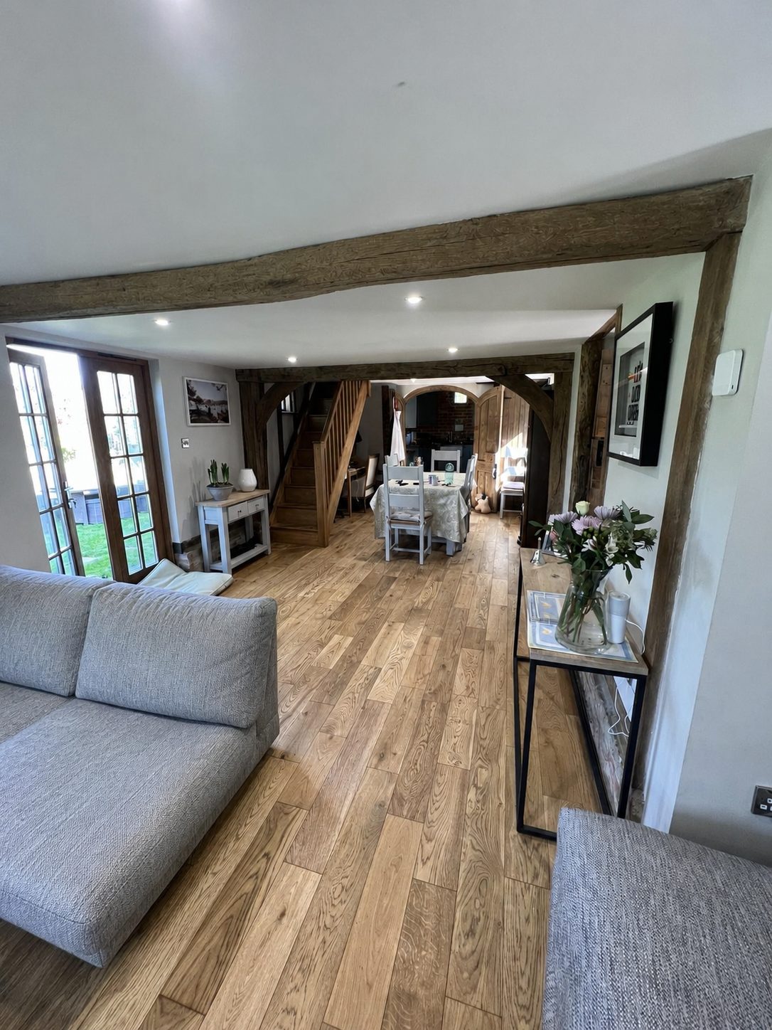 Looking from the kitchen door at The Oast into the sitting and dining space, with a wooden floor, exposed beams, a dining table and chairs and a grey sofa beyond