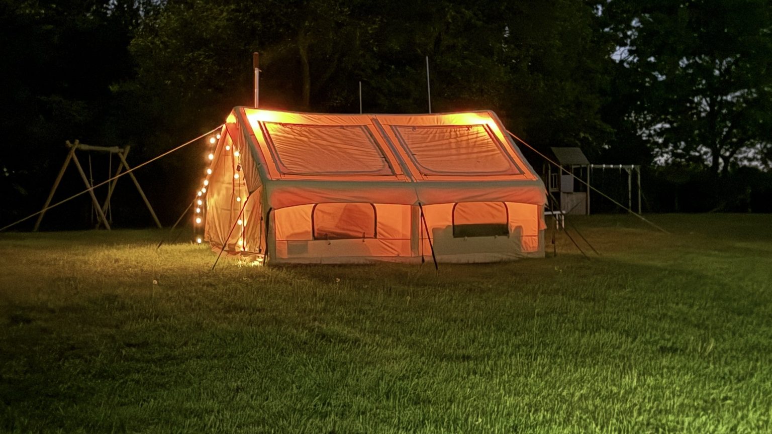 The cream RBM Panda Air Large canvas tent glowing with warm string lights on the lawn at Martins Farm after dark, with trees and the play area behind it.